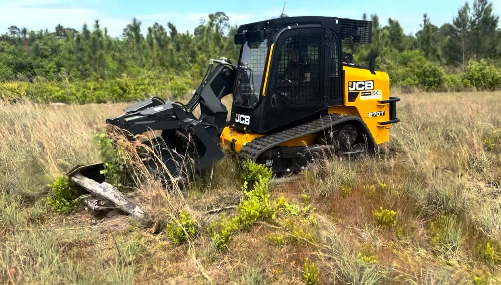 A yellow JCB compact track loader fitted with a mulcher head, cutting through waist-high brush and scrub on an overgrown lot.